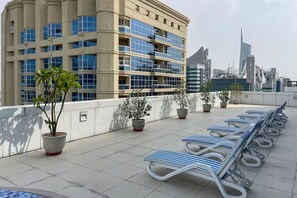 A rooftop terrace with sun loungers, potted plants, and a modern building with large windows. - FRST CLASS |2BR| Urban Living with Stunning Views (JLT)