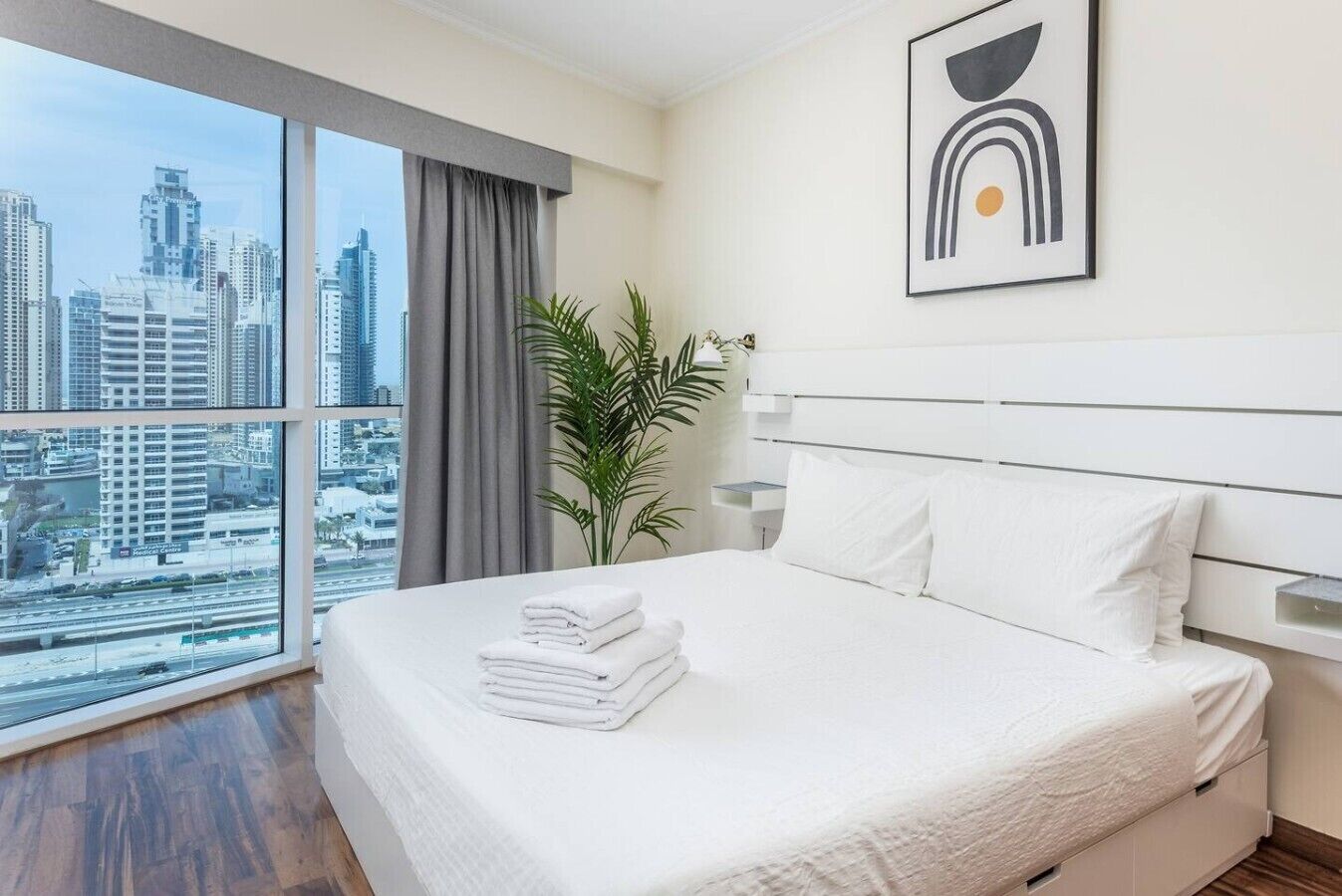Habitación de hotel con un ventanal grande, cama con ropa blanca, cuadro abstracto moderno y una planta en maceta.