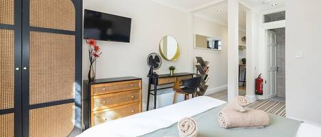 A hotel room with a bed, a desk, a chair, a television, a mirror, and a fire extinguisher.
