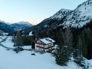 A two-story chalet with a snow-covered roof, surrounded by forested mountains and a clear sky.