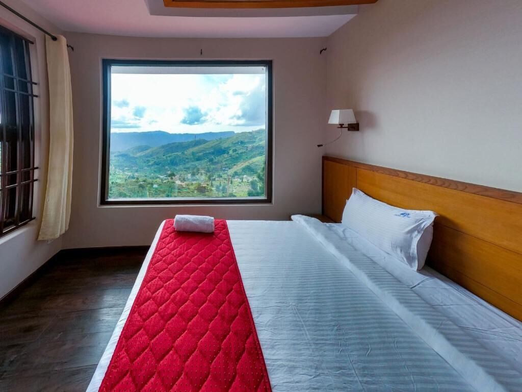 A hotel room with a large window, a bed with a red quilt, and a wooden headboard.