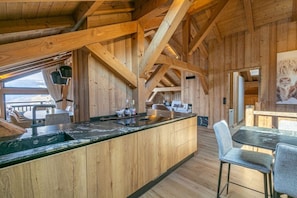A wooden cabin interior with a kitchen island, dining area, and a large window overlooking a deck.