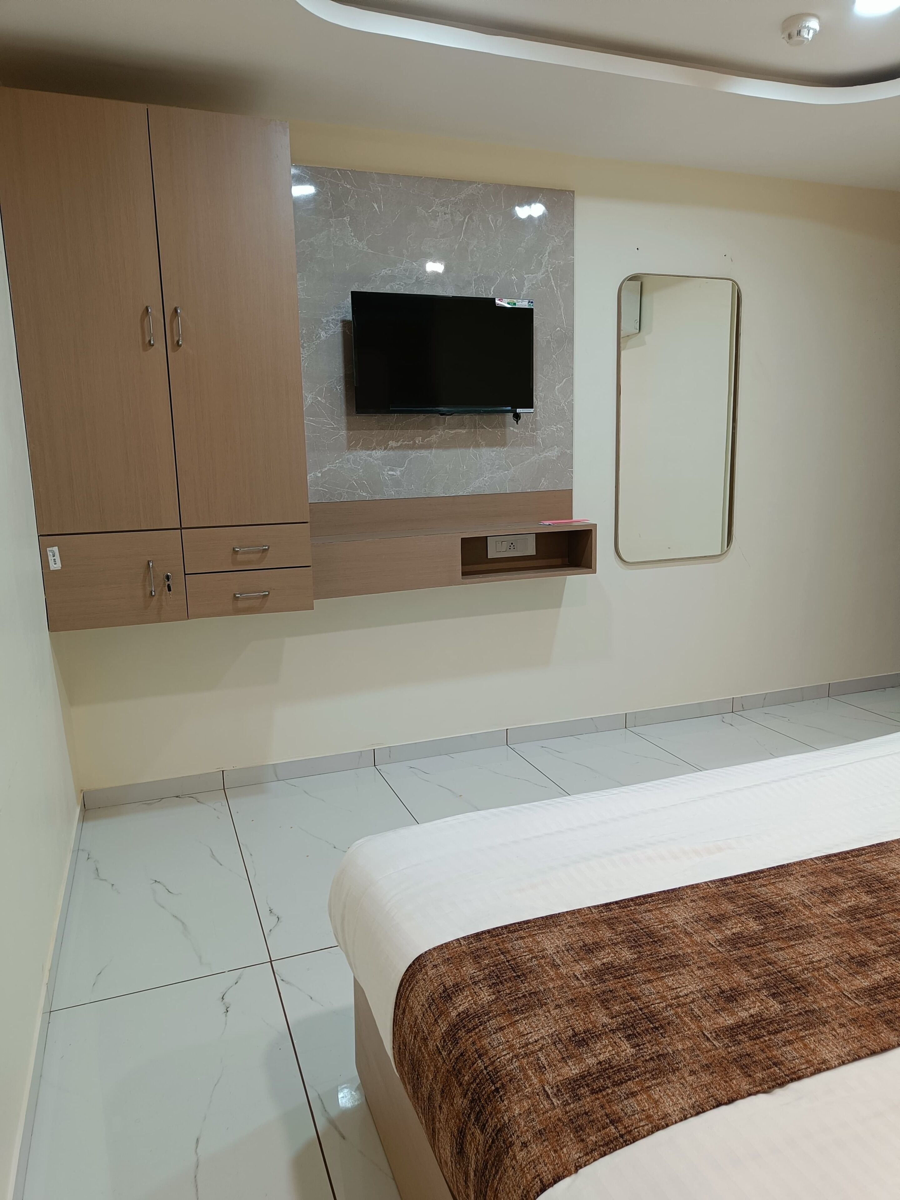 A bedroom with a bed, a TV mounted on a marble wall, a wooden wardrobe, and a mirror.