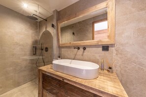 Ein Badezimmer mit einem großen, ovalen Waschbecken, einer Holz-Waschkommode, einer Duschkabine aus Glas und einem Spiegel mit Holzrahmen.