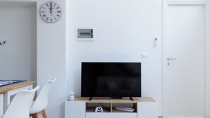 Un salon moderne avec un téléviseur à écran plat posé sur un meuble télé en bois, des chaises blanches, une horloge et une porte.