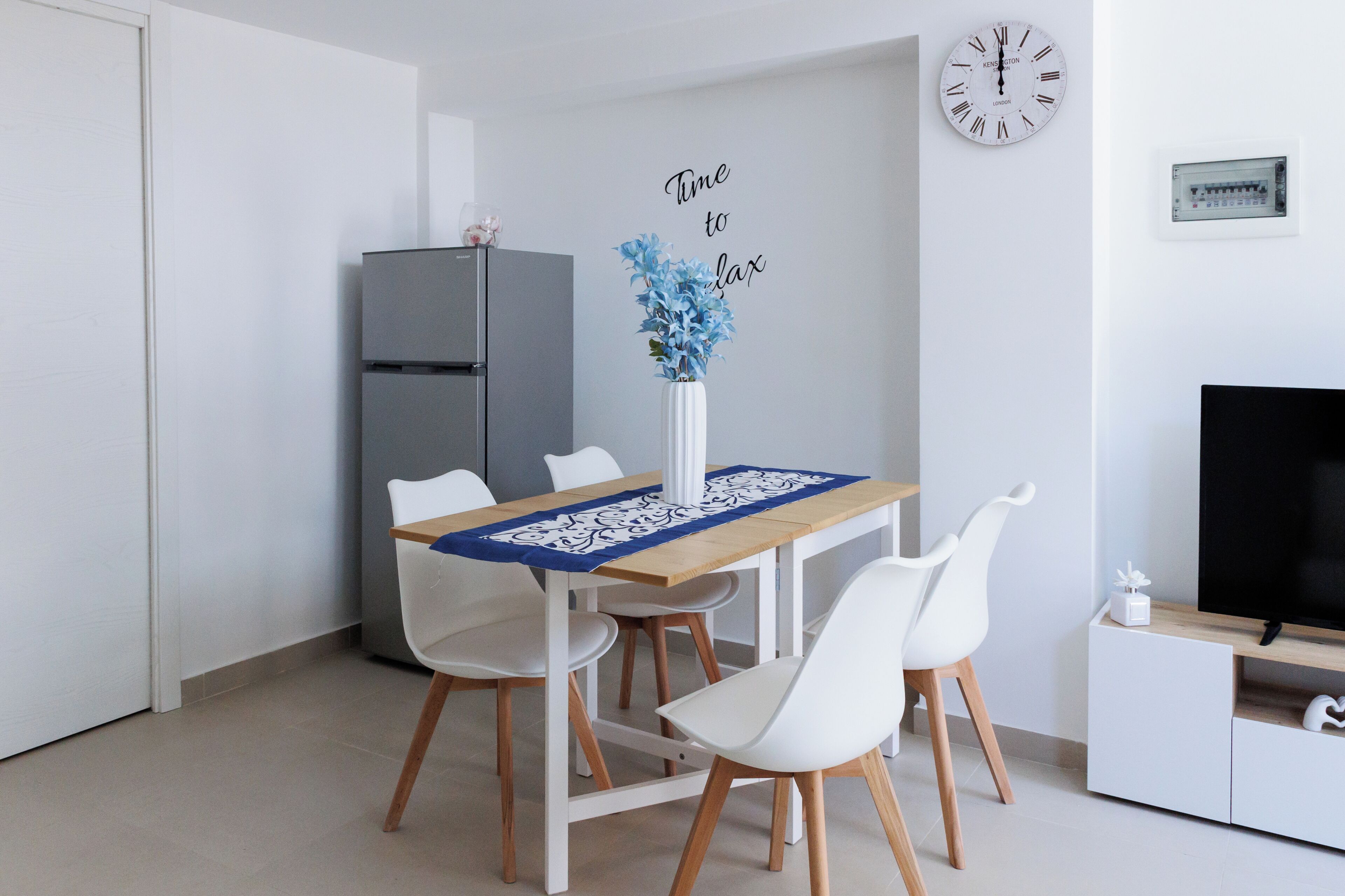 A modern kitchen with a dining table, chairs, a refrigerator, a clock, and a television.