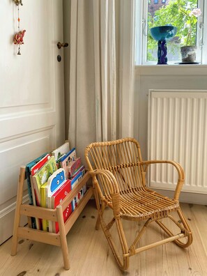 Une pièce avec un fauteuil en osier, une bibliothèque remplie de livres pour enfants, une porte avec un trou de serrure et une fenêtre donnant sur un paysage verdoyant.