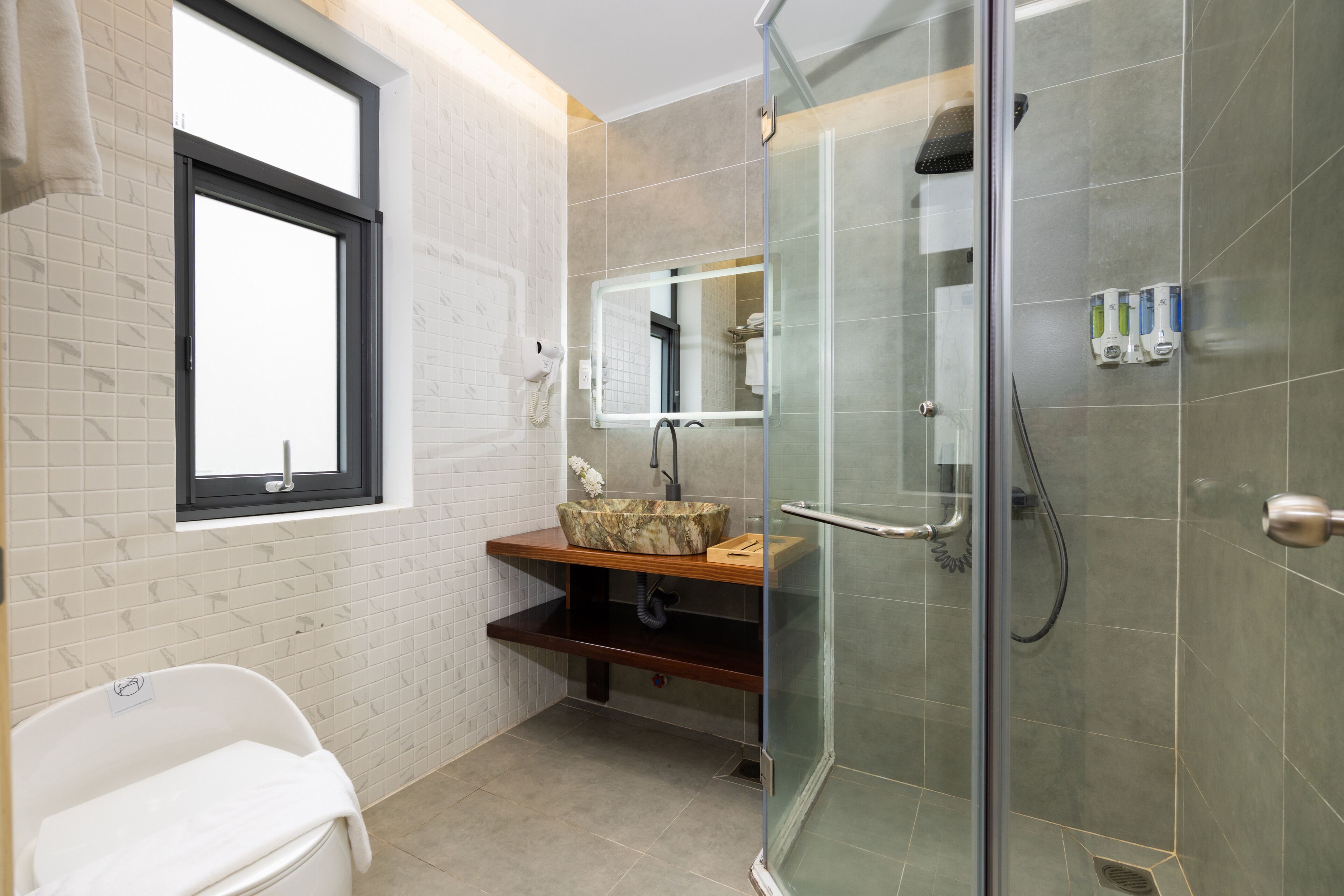 A modern bathroom with a glass shower enclosure, a vanity with a marble basin, and a window above the sink.