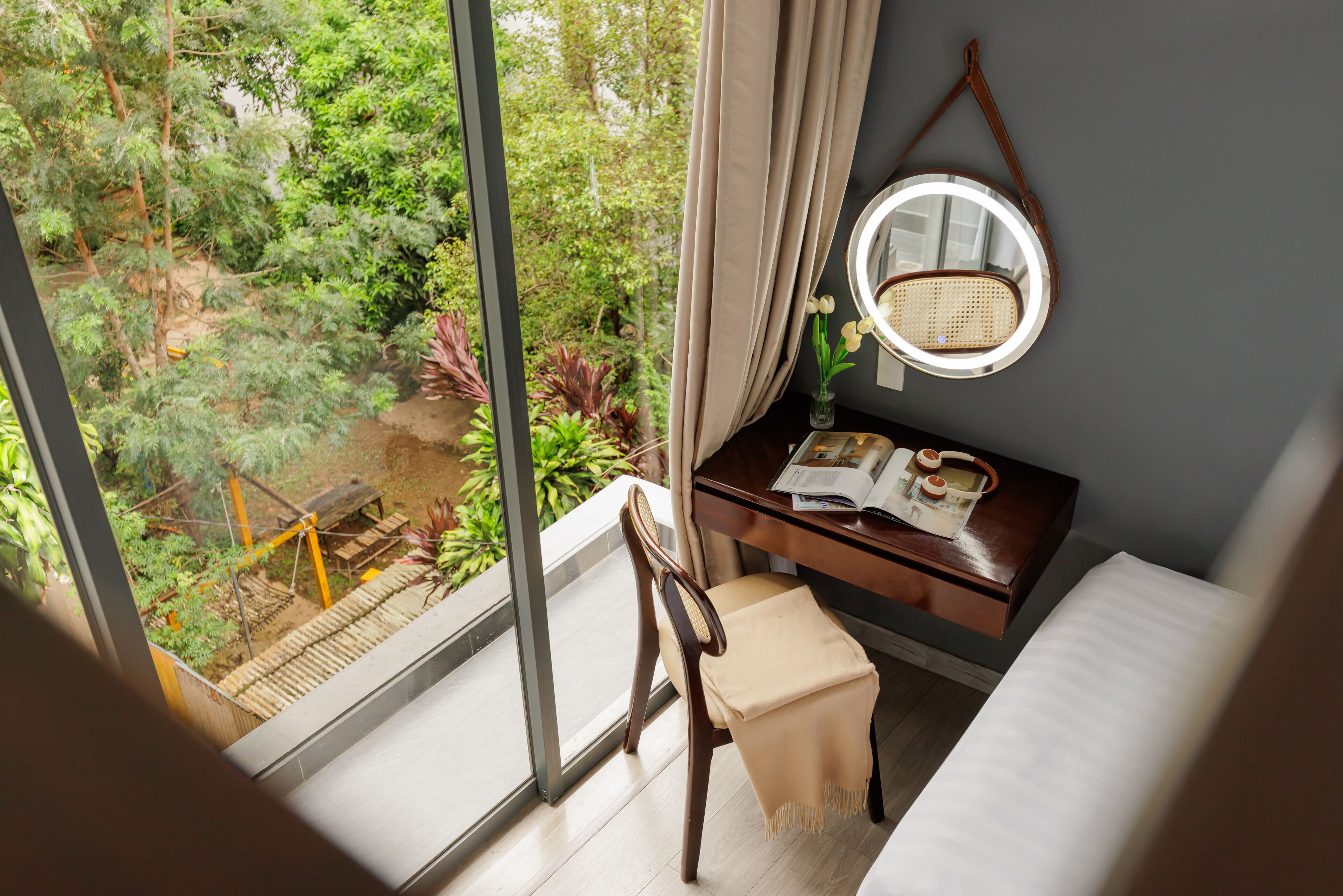A room with a large window, a wooden desk, a chair, a bed, and a round mirror on the wall.