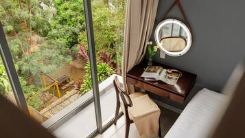 A room with a large window, a wooden desk, a chair, a bed, and a round mirror on the wall.