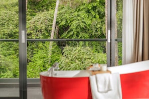 A bathroom with a red bathtub, a white towel, and a view of a dense green forest through large windows. - HAKU Boutique Hotel Da Lat - Ly Tu Trong (Xuan Huong - Da Lat)