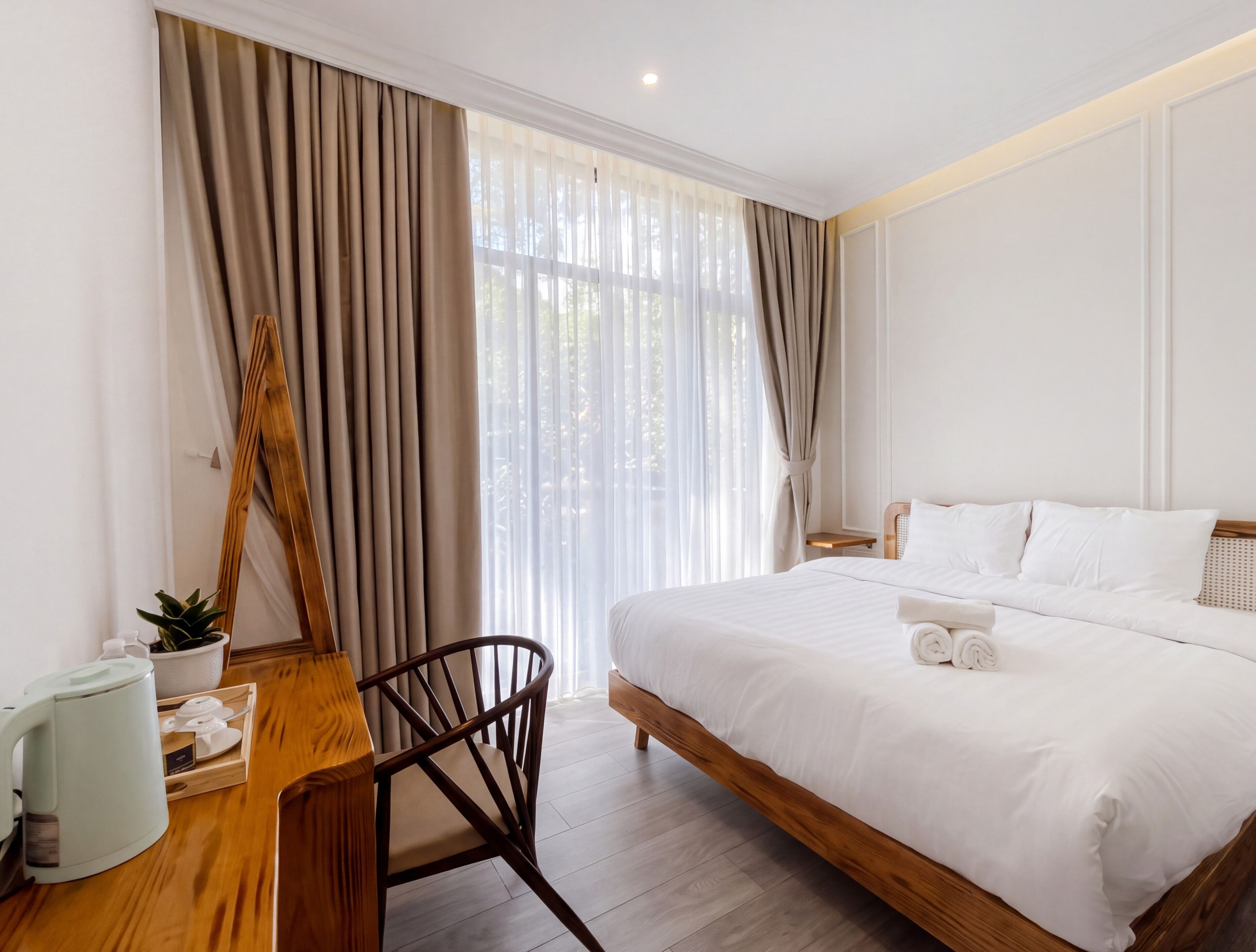 A hotel room with a bed, a wooden desk, a chair, a coffee maker, and a window with curtains.