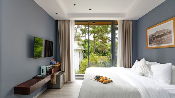 Een moderne hotelkamer met een groot bed, een flatscreen televisie, een bijzettafeltje met lekkernijen, en uitzicht op groen door het raam.