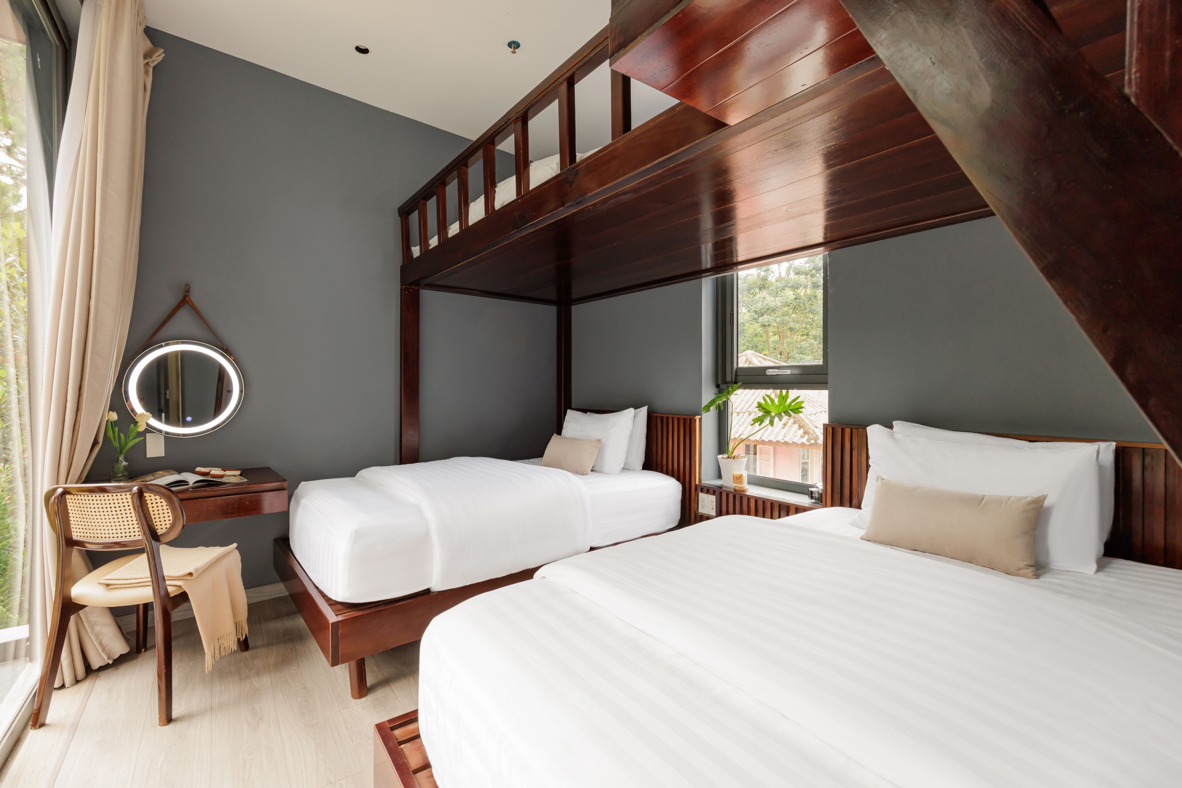 A hotel room with two beds, a desk with a mirror, a chair, and a window with a view of greenery.