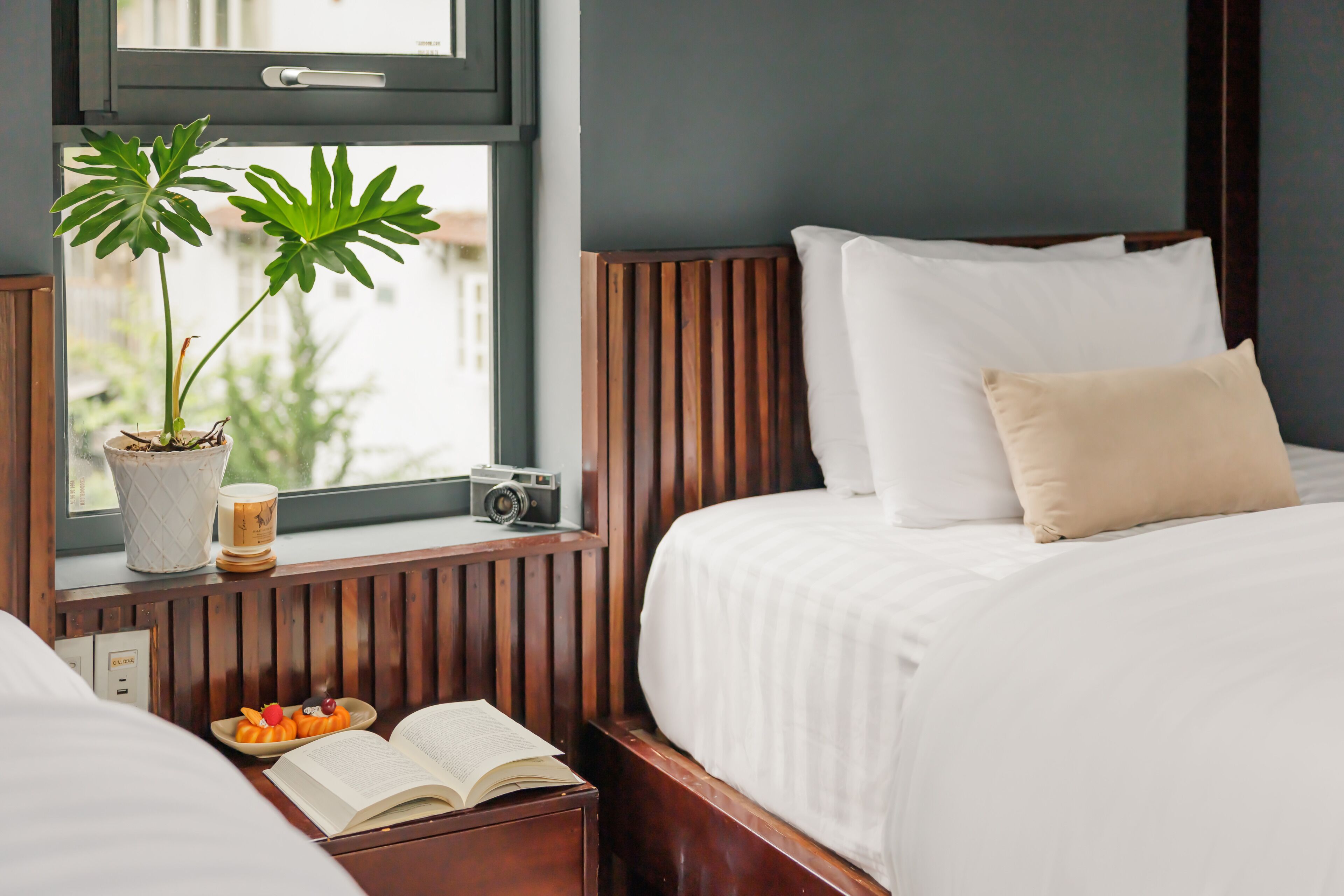 A hotel room with a bed, a bedside table with a book and decorative items, a potted plant, and a window with a view.