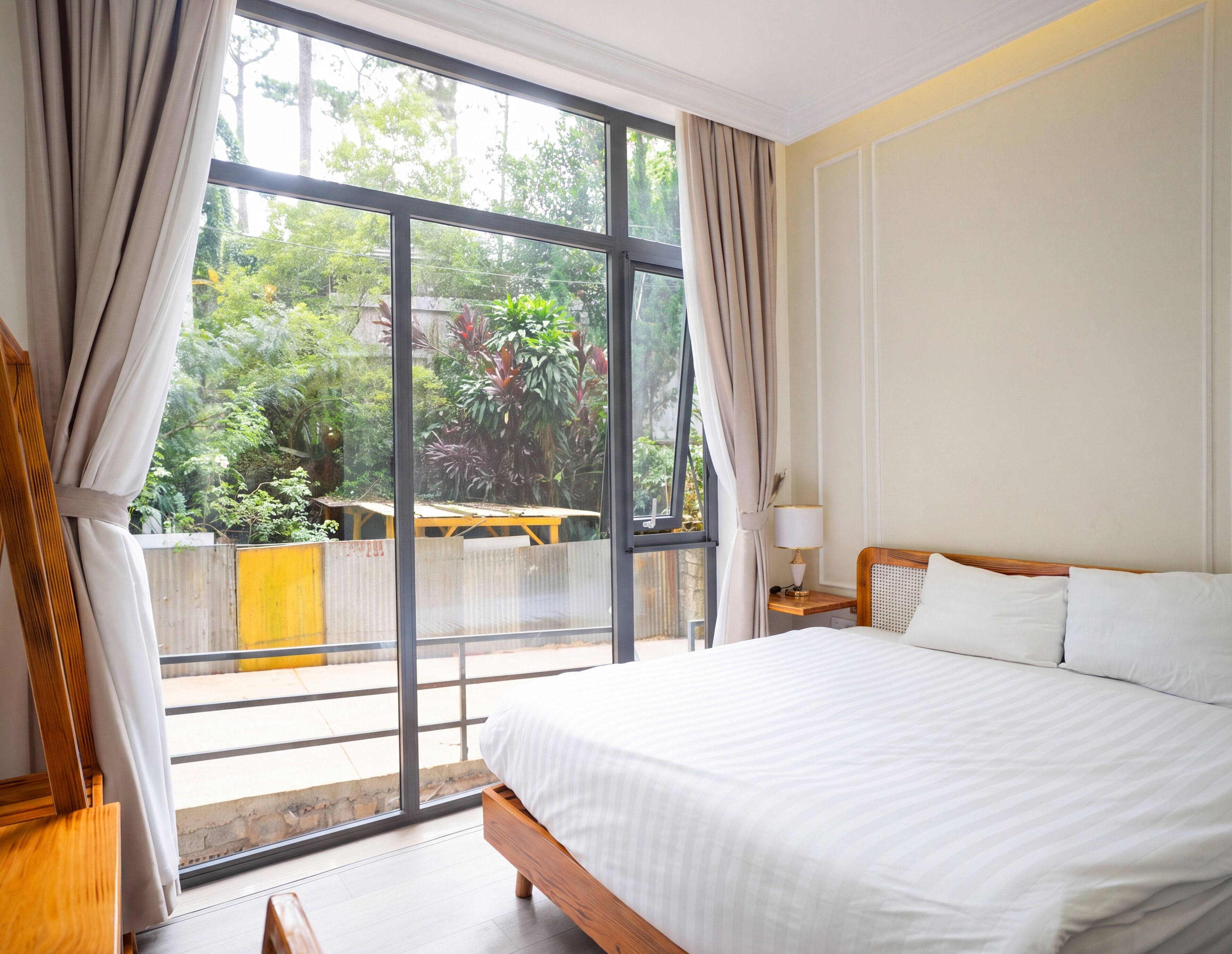 Ein Hotelzimmer mit großem Fenster, einem Bett, einem Nachttisch und Blick auf Grünflächen.