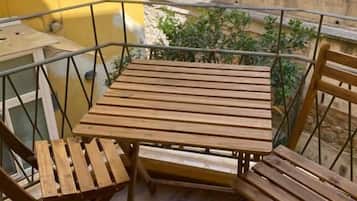 A balcony with a wooden table and chairs, a yellow wall, and a view of a street with cars and greenery.