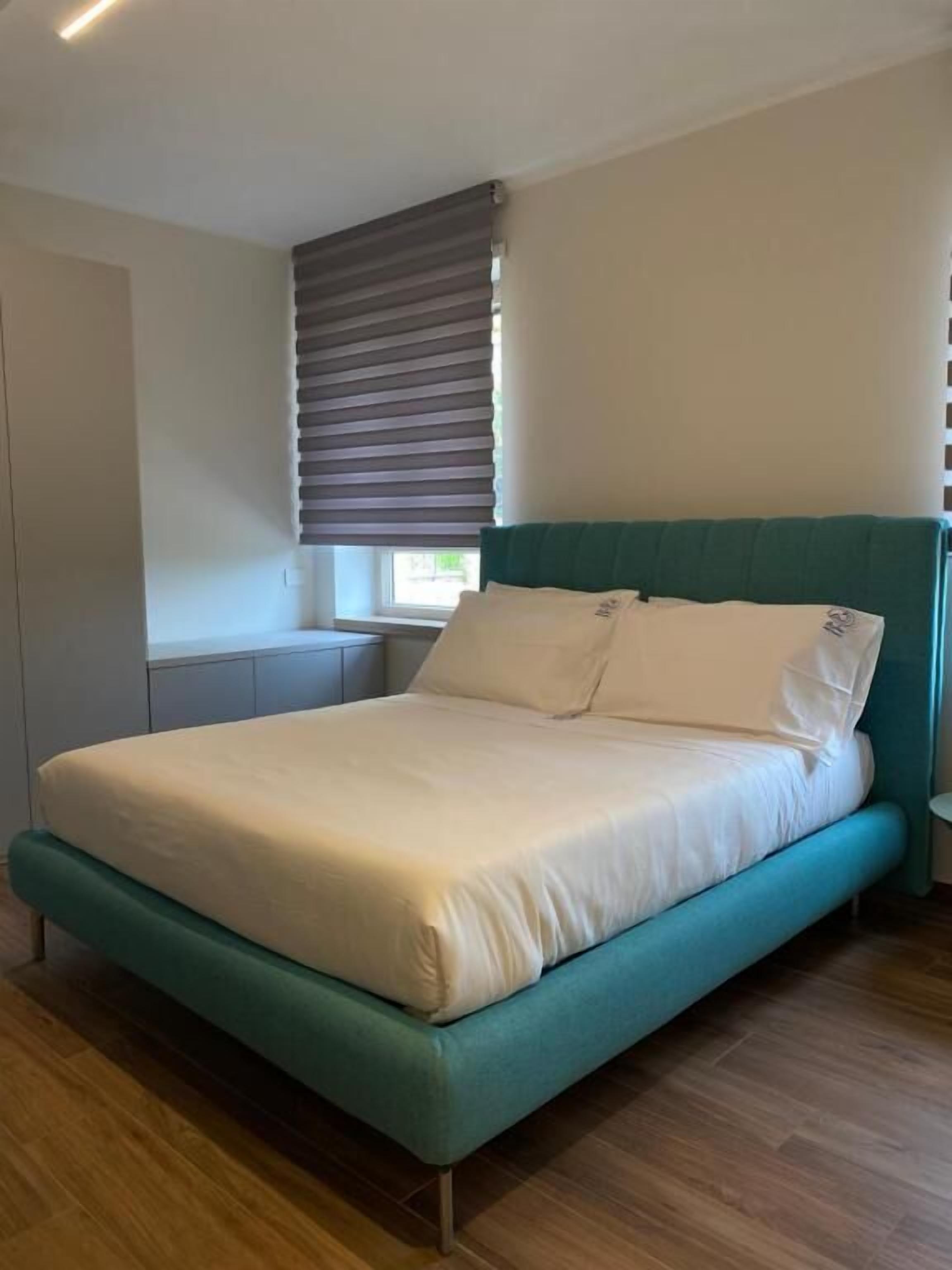 A modern bedroom with a teal bed, white bedding, a window with blinds, and a wooden floor.