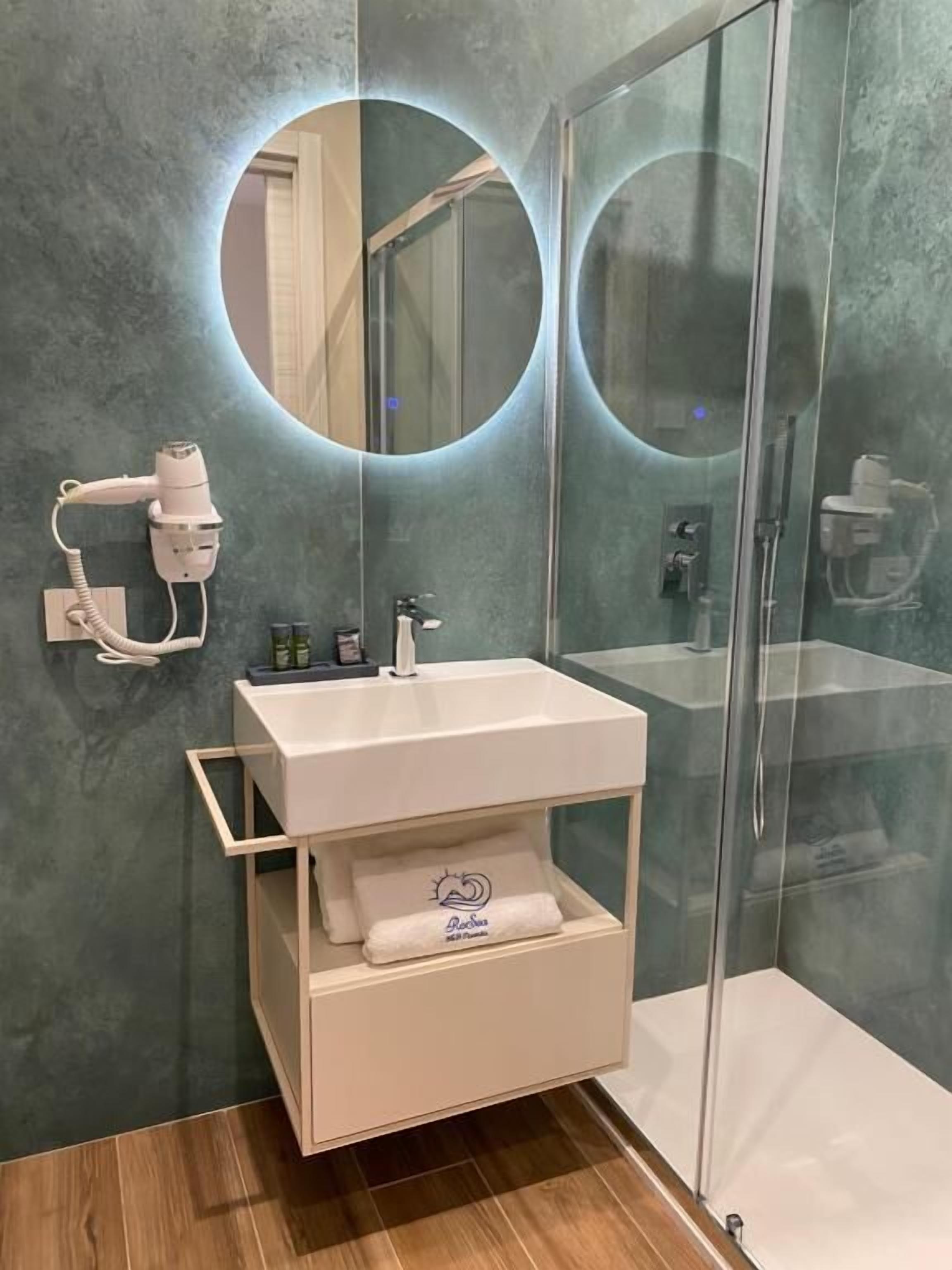 A bathroom with a glass shower enclosure, a white sink with a towel rack, and a mirror with a circular light.