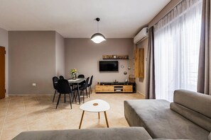A modern living room with a sofa, dining table, chairs, a TV, and a TV stand. - Coastal Living Retreat | 2BR in Lungomare by PikHost (Vlorë)
