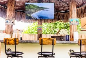 Eine Barbereich mit einem Fernsehbildschirm, der Blick aufs Meer zeigt, Holzsessel und einem Schild, das zur Terrasse einlädt.