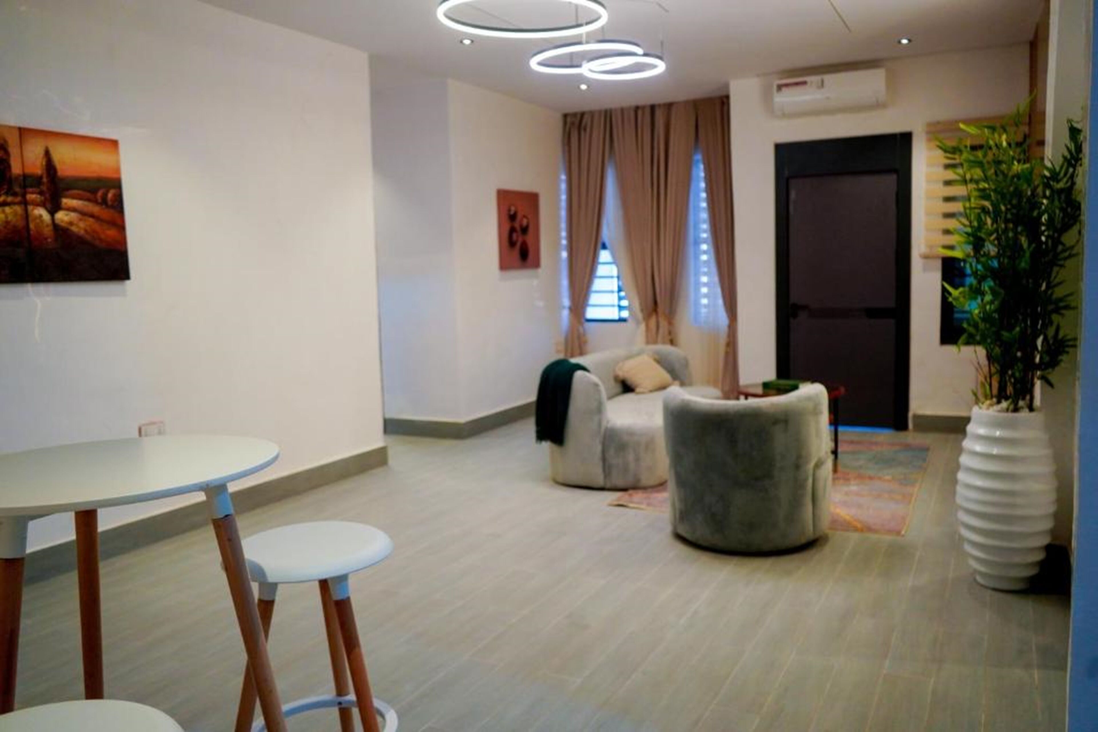 A modern hotel lobby with a round table, two bar stools, a grey sofa, a potted plant, and a painting on the wall.