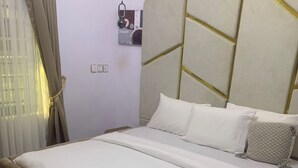 Uma cama com cabeceira bege, roupa de cama branca e uma pequena mesa de cabeceira com um abajur.
