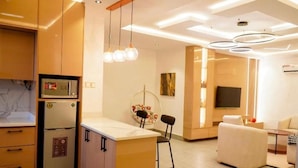 Uma cozinha moderna com eletrodomésticos embutidos, uma área de refeições com um banquinho de bar e uma sala de estar com sofá e televisão.
