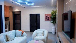 Quarto de hotel moderno com sofá, poltrona, mesa redonda, TV de tela plana e uma planta.
