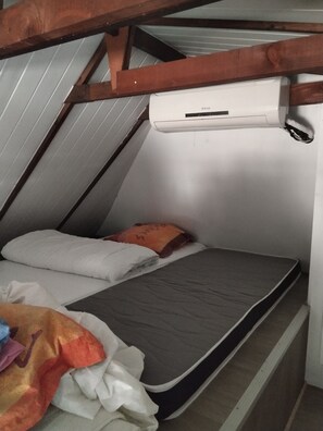 A small, slanted room with a single bed, a pillow, a blanket, and a wall-mounted air conditioning unit.