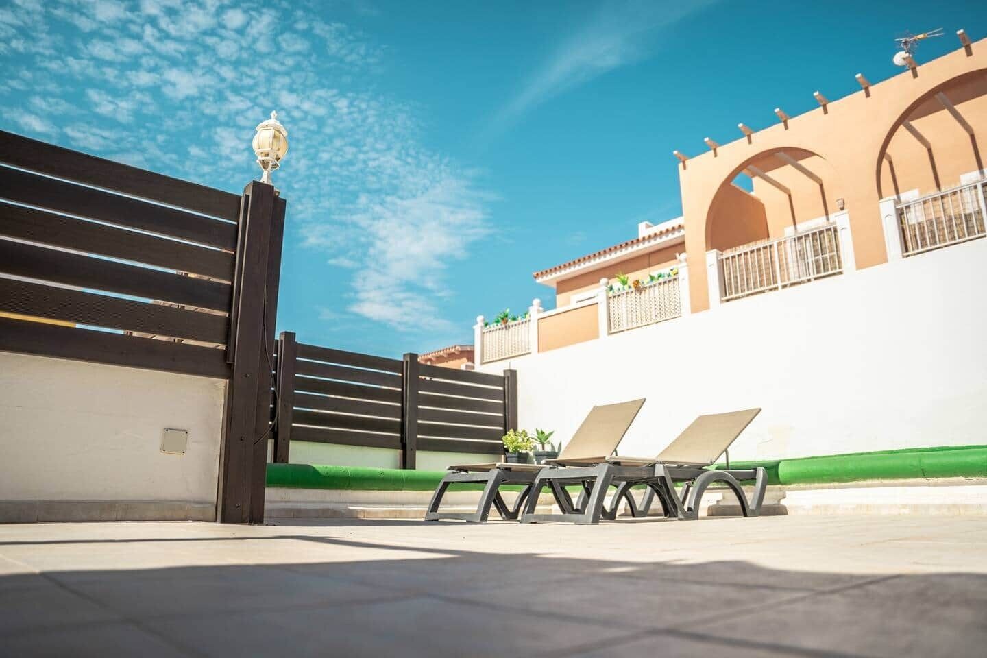 A gated entrance with a lamp post, a white wall with a green base, and two lounge chairs.