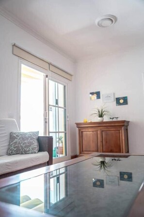 Een kamer met een tafel met glazen blad, een bank met opvallende kussens, een houten kast en een schuifpui naar een buitenruimte.