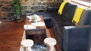 A cozy living area with a stone wall, a sofa with a yellow throw, a coffee table with books, and a potted plant.