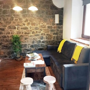 Ein gemütlicher Wohnbereich mit einer Steinwand, einem Sofa mit gelber Decke, einem Couchtisch mit Büchern und einem Blumentopf.