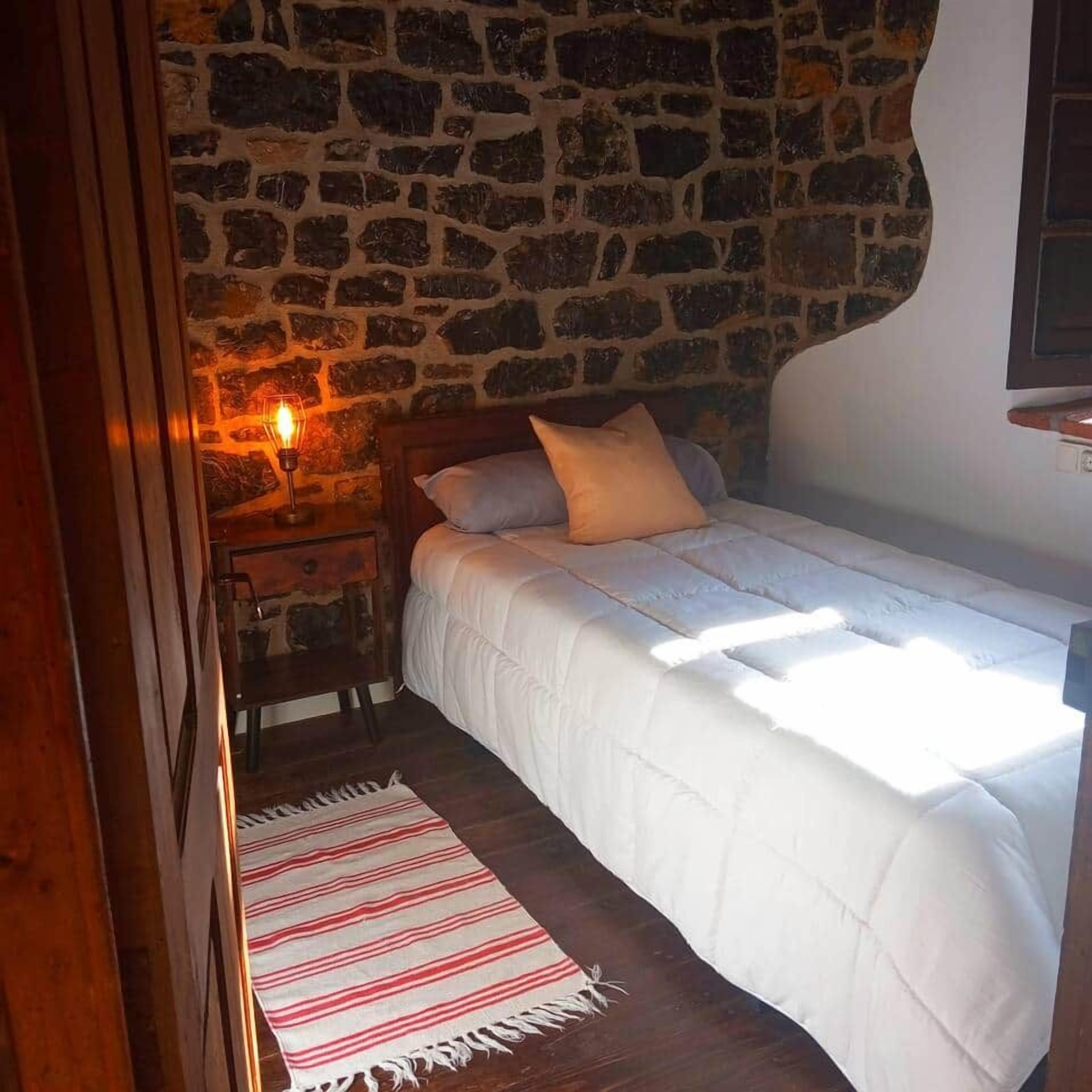 A bedroom with a stone wall, a bed with white bedding, a red and white striped rug, and a nightstand with a lamp.