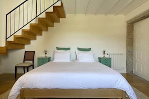 Ein Schlafzimmer mit einer Holztreppe, einem Bett mit weißer Bettwäsche, zwei Nachttischen mit Lampen, einem Sessel und einem Heizkörper.