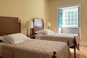 A room with two wooden beds, each with a floral-patterned bedspread and a wooden headboard. There are bedside tables with lamps and a window with a view of greenery. - Family house with pool at Quinta da Agrela (Portugal) (Vade (São Pedro))