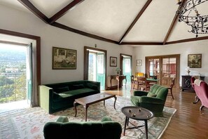 Ein geräumiges Wohnzimmer mit einem grünen Sofa, einem Couchtisch und einem gemusterten Teppich. Zwei Türen führen in andere Räume, und eine Chandelüster hängt von der Decke.