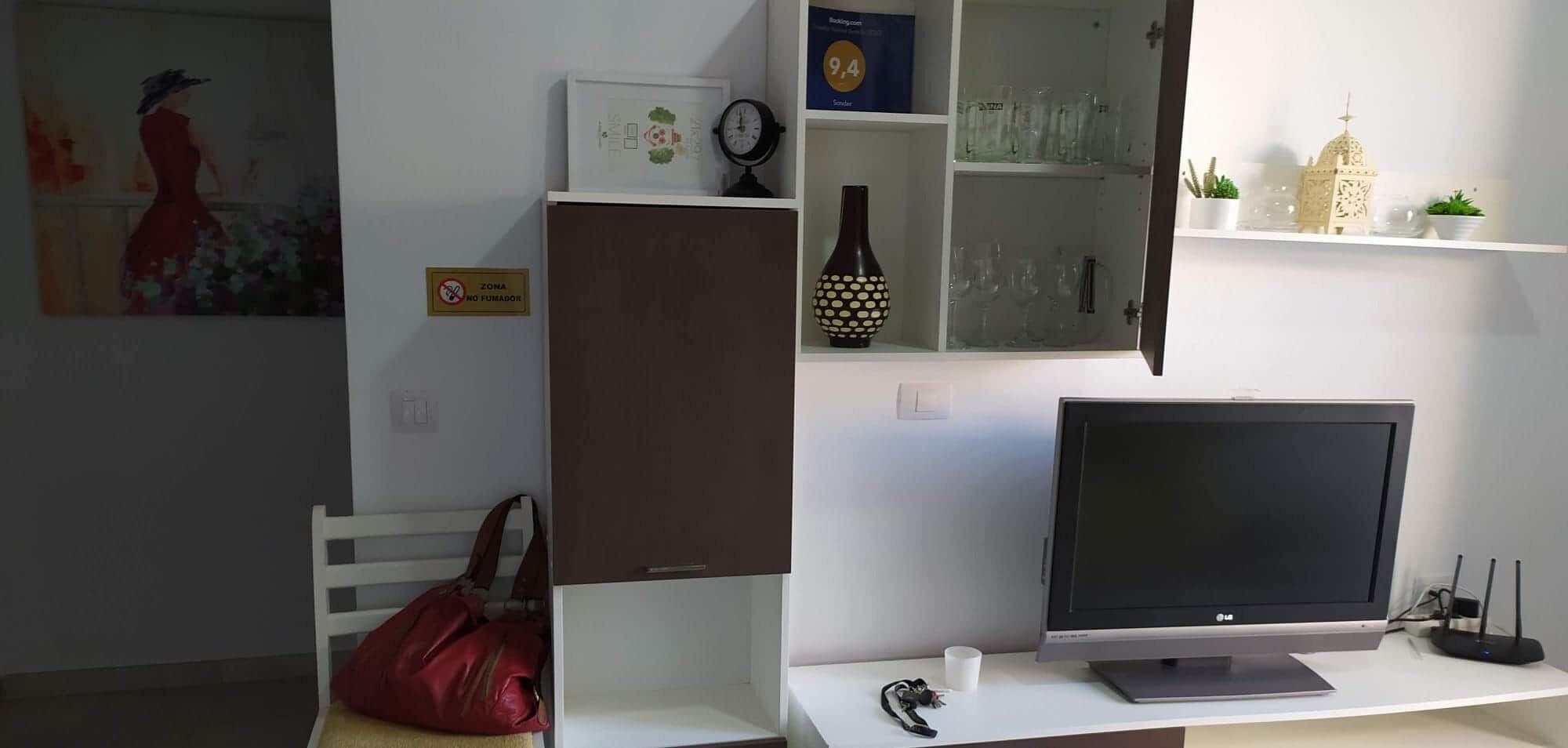 Une pièce comprenant un bureau, un écran d’ordinateur, un sac rouge, une horloge et une étagère avec des objets décoratifs.