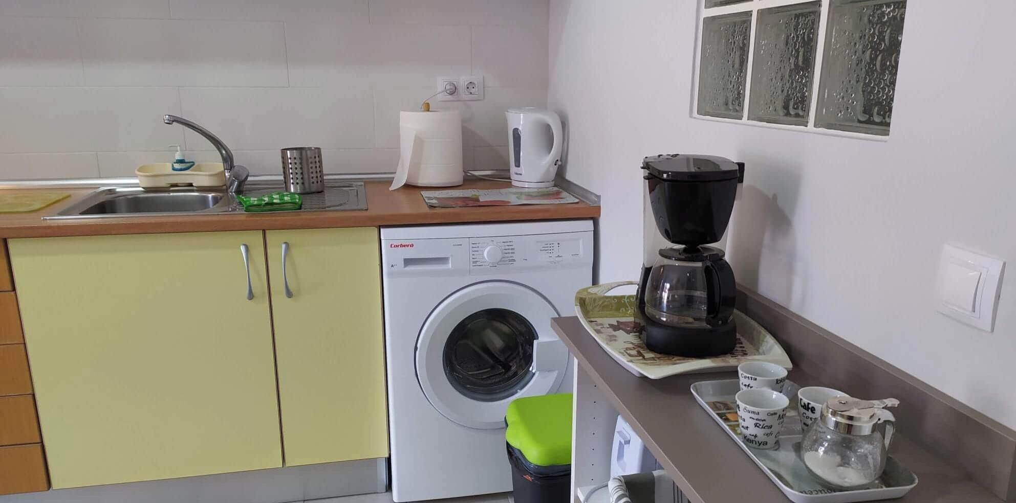 Een kleine keuken met een wasmachine, koffiezetapparaat en een prullenbak.
