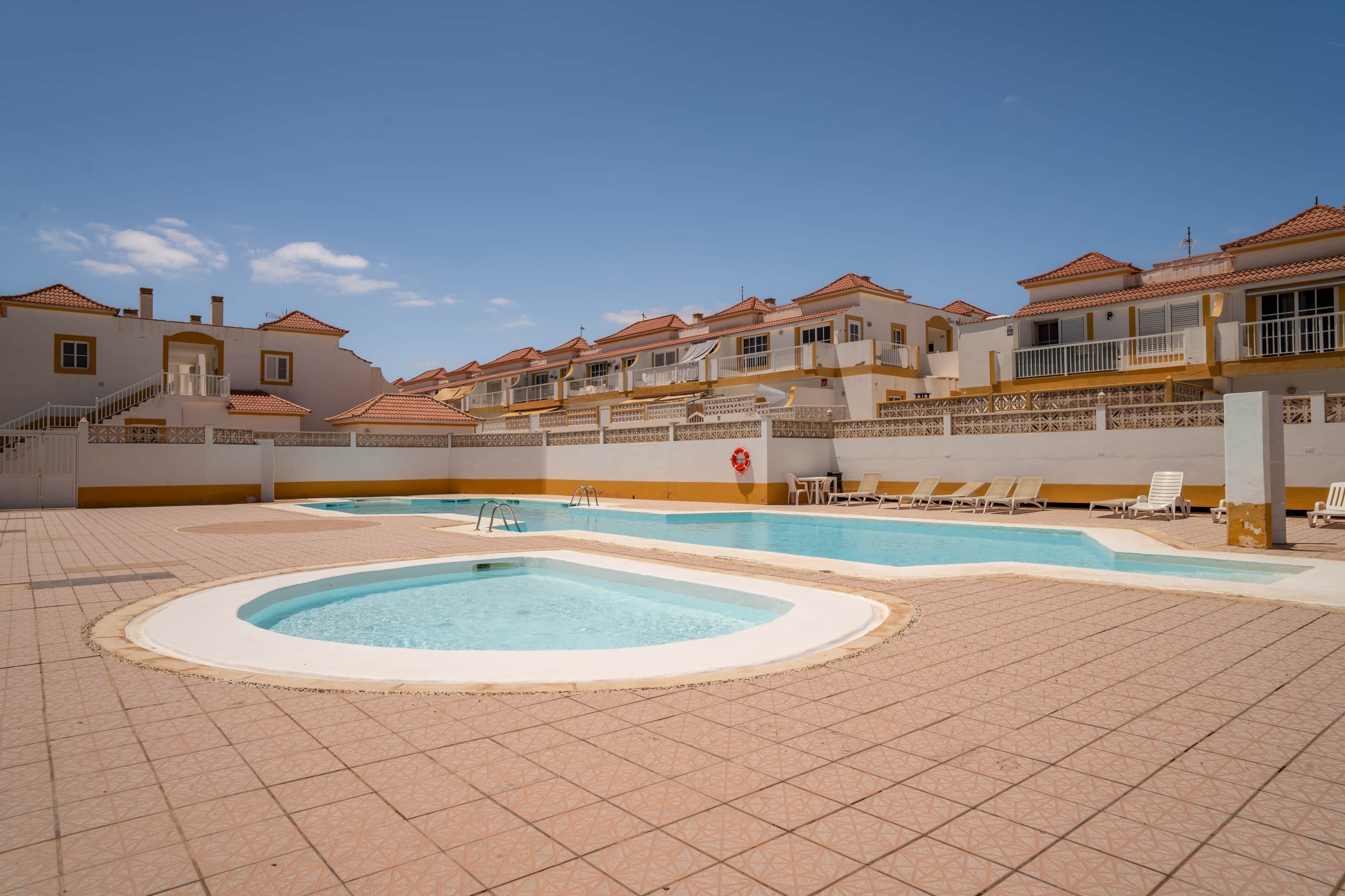 Zona de piscina con suelo de baldosas, sillas de descanso y un edificio con varios balcones.