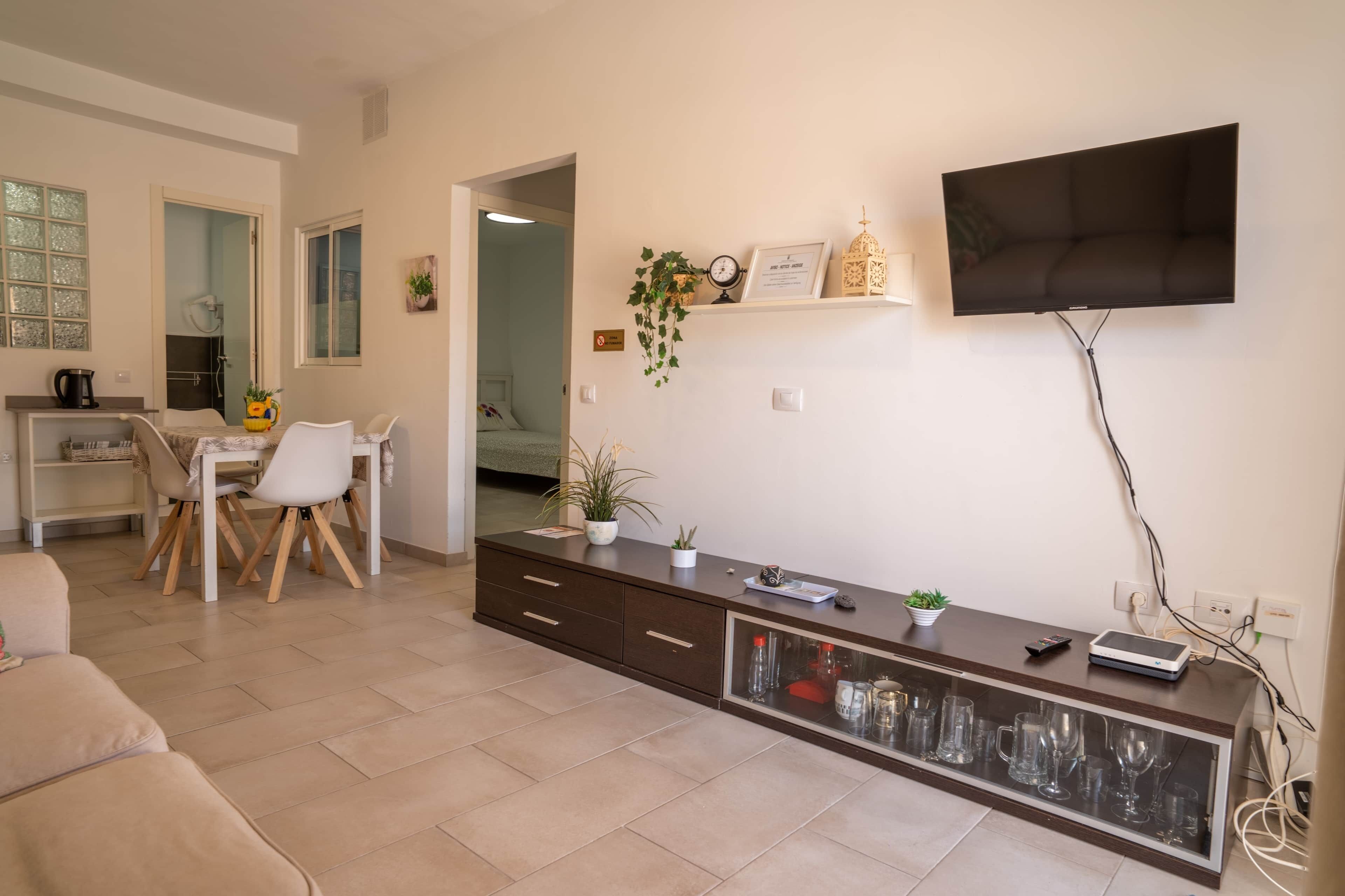 Una sala de estar moderna con un televisor de pantalla plana, un comedor con mesa y sillas, y una cocina con hervidor.