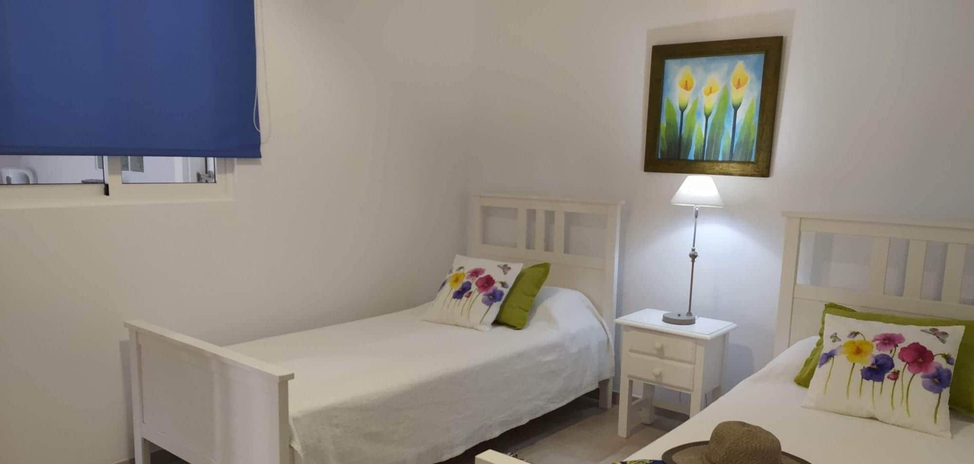 Un dormitorio con dos camas individuales, una mesita de noche con lámpara, un cuadro colgado en la pared y una ventana con persiana azul.