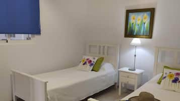 Un dormitorio con dos camas individuales, una mesita de noche con lámpara, un cuadro colgado en la pared y una ventana con persiana azul.