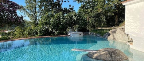 A clear swimming pool with a rock feature, surrounded by trees and a white building.
