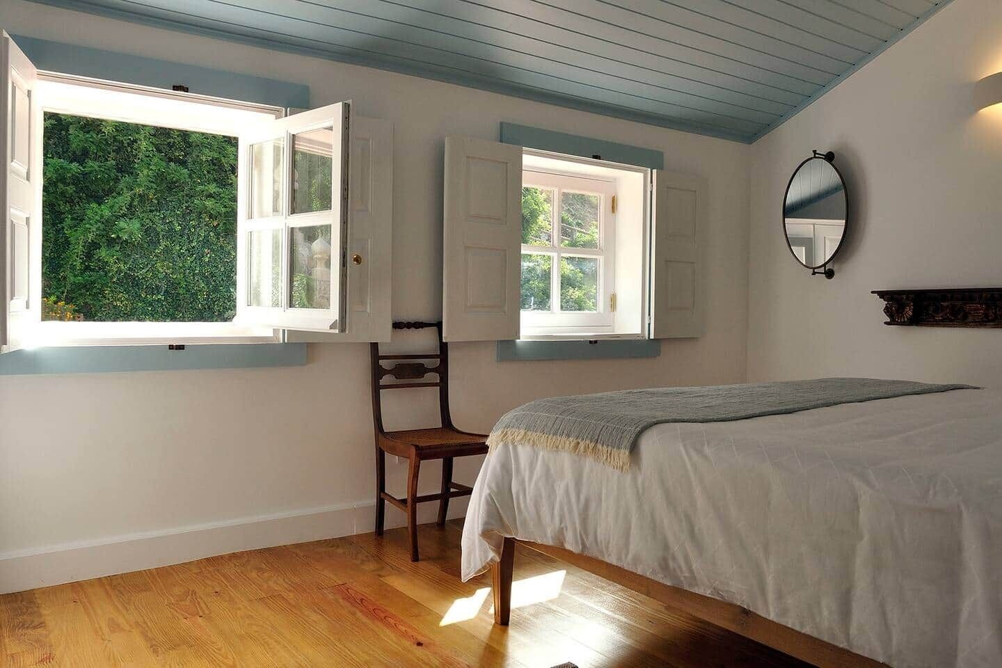 A bedroom with a bed, two windows with open shutters, a chair, and a mirror on the wall.