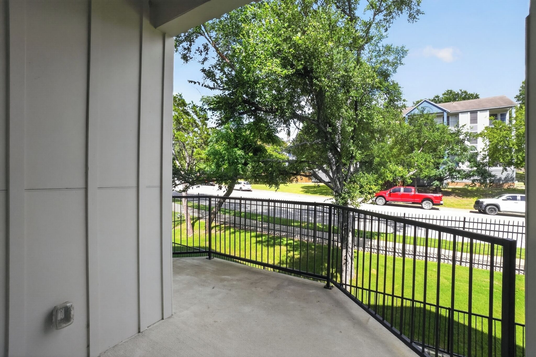 A balcony with a black metal railing, a concrete floor, and a view of a street with parked cars and a tree.