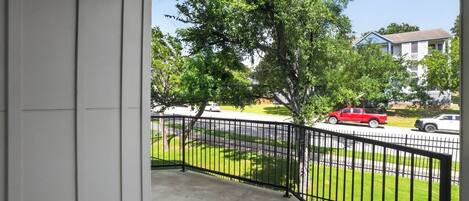 A balcony with a black metal railing, a concrete floor, and a view of a street with parked cars and a tree.
