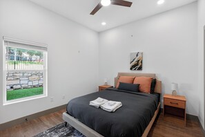 Ein Schlafzimmer mit einem Bett, Nachttischen, einem Deckenventilator, einem Fenster mit Blick auf eine Steinmauer und Gras sowie einem Bild an der Wand.