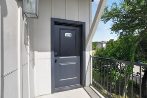 A dark-colored door with a number plate, a balcony with a metal railing, and a tree in the background. - Elegant 2-bedroom with upscale finishes throughout. (Austin)